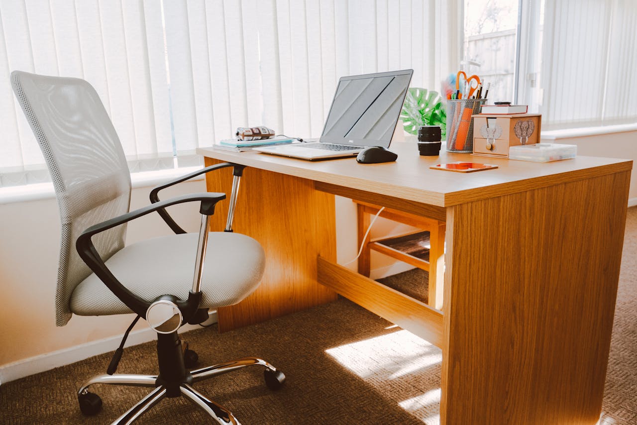 A modern office workspace featuring a wooden desk, chair, laptop, and bright natural light.